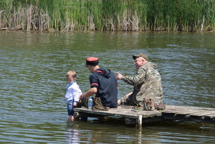 молодой казак держит сына на деревянной пристани на реке Кирпили, рядом мужчина в камуфляжной форме