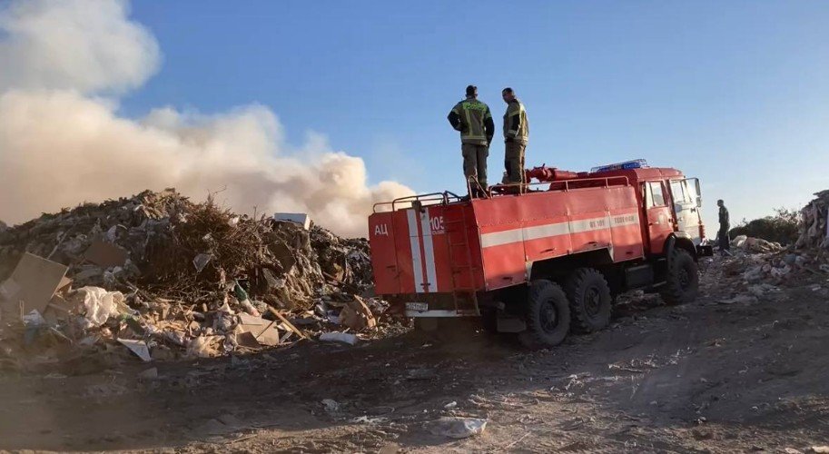 пожарные стоят на пожарной машине и смотрят на дымящийся мусорный полигон