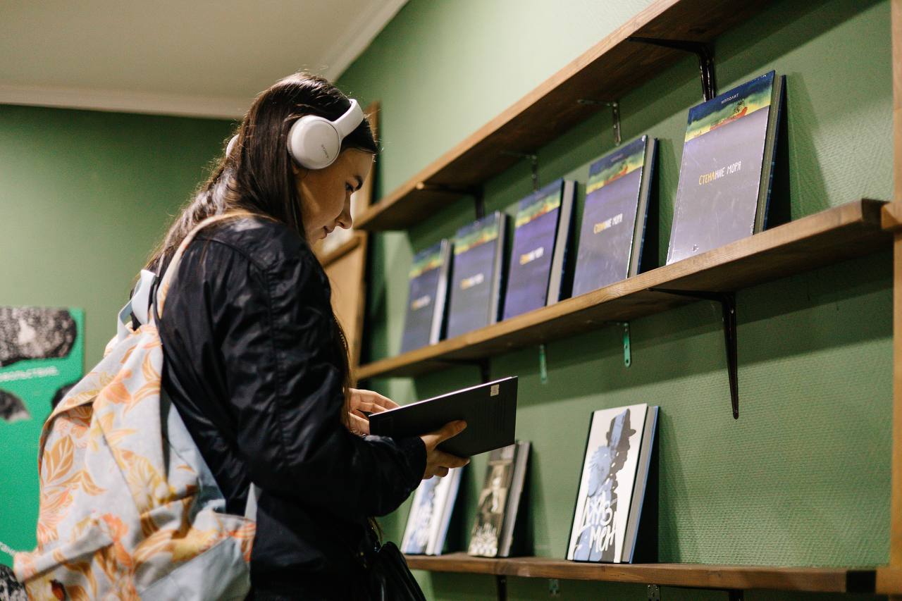 девушка в наушниках листает книгу перед книжными полками в магазине